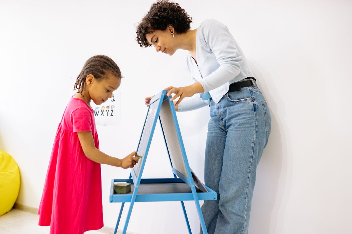 A teacher guiding a young girl as she draws on a chalkboard easel during a session